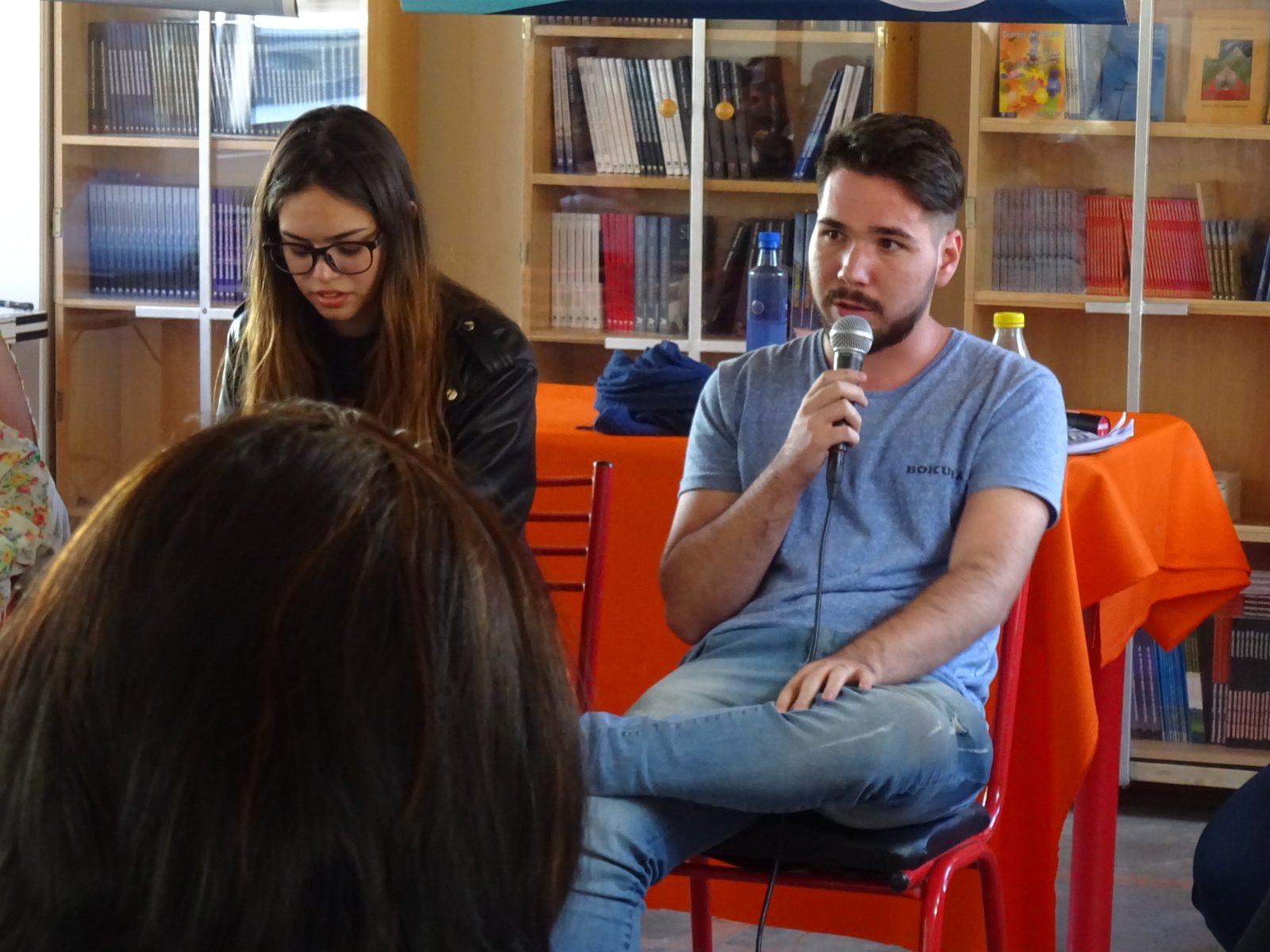 Charla de COPEHU: una persona habla con micrófono ante el público, con una biblioteca de fondo.